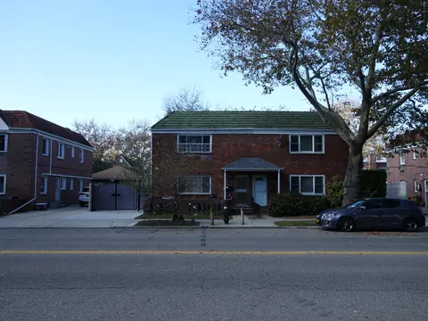 Two-story brick houses with lawns and trees on a wide residential street in eastern Queens