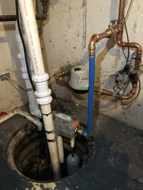 A sump pump standing in its pit in a basement floor, with its discharge pipe and water lines beside it