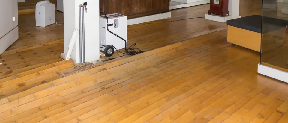 A dehumidifier running beside a pillar on a water-damaged wood floor, with floorboards lifted at its base