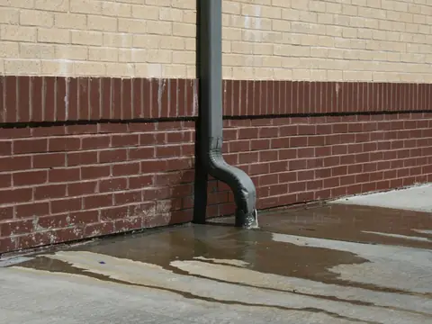 A downspout emptying rainwater onto the pavement beside a brick wall